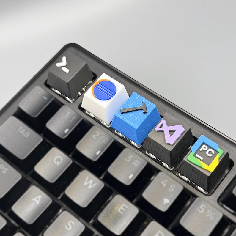 以下が含まれることがあります： カスタムキーキャップが付いた黒いキーボードのクローズアップ。キーキャップには、ハンマー、線のある円、PCアイコンなど、さまざまなカラフルなデザインとシンボルが表示されています。キーボードには、白い文字が入った灰色のキーがあります。