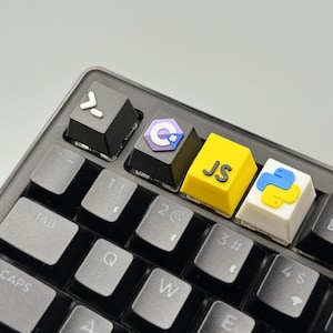Puede incluir: Primer plano de un teclado negro con teclas personalizadas. Las teclas presentan s&iacute;mbolos de lenguajes de programaci&oacute;n: una tecla negra con una flecha hacia la derecha, una tecla con el logotipo de C#, una tecla amarilla con "JS" y una tecla blanca con el logotipo de Python.