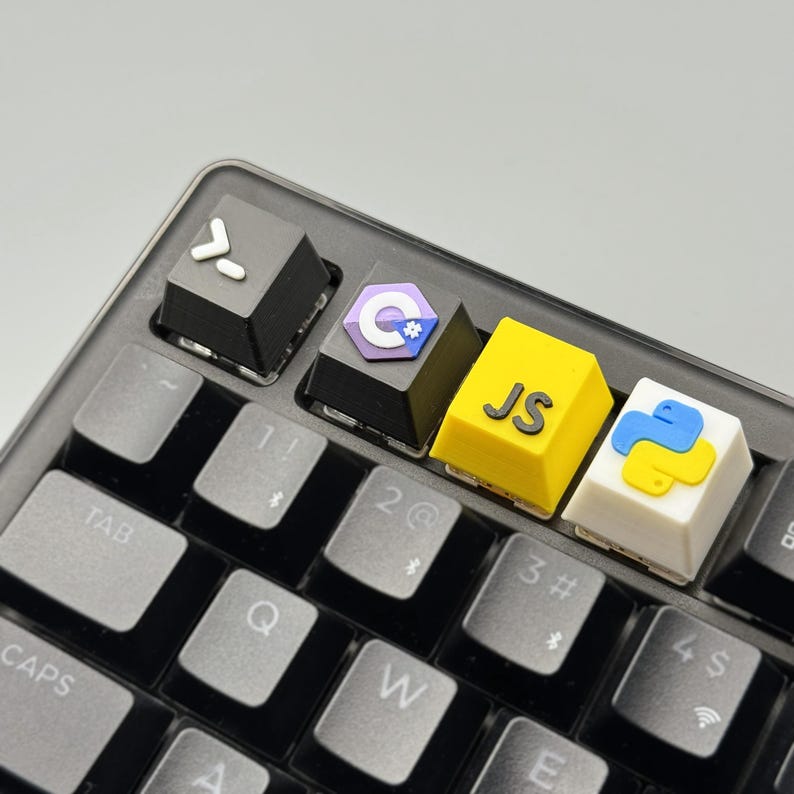 Puede incluir: Primer plano de un teclado negro con teclas personalizadas. Las teclas presentan s&iacute;mbolos de lenguajes de programaci&oacute;n: una tecla negra con una flecha hacia la derecha, una tecla con el logotipo de C#, una tecla amarilla con "JS" y una tecla blanca con el logotipo de Python.
