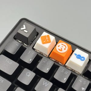 Puede incluir: Primer plano de un teclado negro con teclas personalizadas. Las teclas presentan varios s&iacute;mbolos y logotipos en blanco, naranja y azul. El teclado incluye las teclas Tab, Q, W y otras.