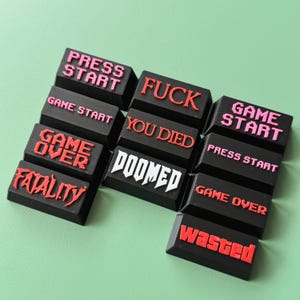 May include: A set of black keycaps with red and white text. The keycaps feature phrases like "Press Start", "Game Over", "You Died", "Doomed", "Fatality", "Wasted", and "Fuck".