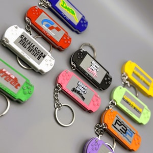 Puede incluir: Una colección de llaveros PSP coloridos. Los llaveros son de varios colores, incluyendo rojo, rosa, verde y negro. Cada llavero presenta un título de juego en la pantalla, como "Grand Theft Auto" y "Crisis Core Final Fantasy VII".