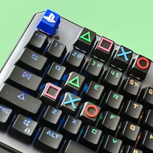 May include: A black keyboard with blue and white LED lights. The keyboard has custom keycaps with PlayStation controller symbols, including a triangle, square, circle, and X. The keycaps are in various colors, including green, pink, blue, and red.