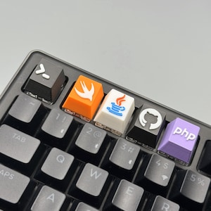 Puede incluir: Primer plano de un teclado mec&aacute;nico con teclas personalizadas. Las teclas presentan logotipos de lenguajes de programaci&oacute;n, como Swift, Java, GitHub y PHP. El teclado tiene un marco negro y teclas grises.