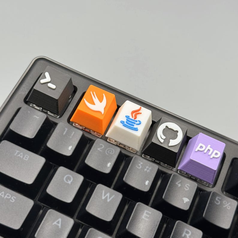Puede incluir: Primer plano de un teclado mec&aacute;nico con teclas personalizadas. Las teclas presentan logotipos de lenguajes de programaci&oacute;n, como Swift, Java, GitHub y PHP. El teclado tiene un marco negro y teclas grises.