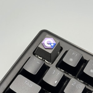 Puede incluir: Una tecla personalizada con un dise&ntilde;o p&uacute;rpura, blanco y azul, instalada en un teclado mec&aacute;nico negro. La tecla presenta una letra "C" estilizada y un icono de engranaje. El teclado tiene teclas grises y est&aacute; en &aacute;ngulo.