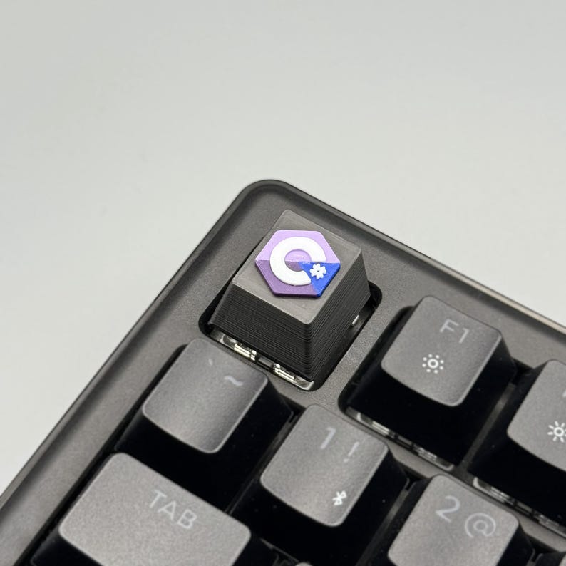 Puede incluir: Una tecla personalizada con un dise&ntilde;o p&uacute;rpura, blanco y azul, instalada en un teclado mec&aacute;nico negro. La tecla presenta una letra "C" estilizada y un icono de engranaje. El teclado tiene teclas grises y est&aacute; en &aacute;ngulo.