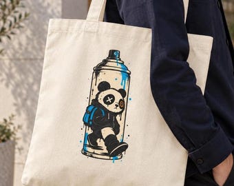 May include: A cream-colored canvas tote bag featuring a graphic print of a spray paint can. Inside the can is a stylized panda wearing a blue backpack, with one eye marked by an X and the other by a circular orange detail, accented by blue paint splatters.