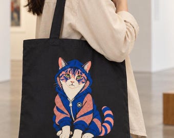 May include: A black tote bag featuring a graphic print of a cat wearing a blue and orange zip-up hoodie. The cat has green eyes and a striped tail, and the hoodie includes a small circular logo on the chest. The bag has two black shoulder straps.