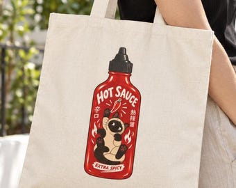 May include: A beige canvas tote bag featuring a graphic of a red hot sauce bottle with a black cap. The label displays a Siamese cat, flames, a chili pepper icon, and the text "HOT SAUCE", "EXTRA SPICY", "辛口", and "熱辣醬".