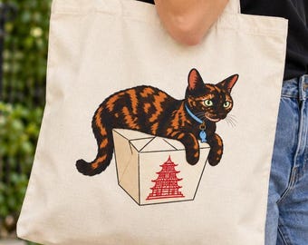 May include: A beige canvas tote bag featuring a graphic illustration of a tortoiseshell cat resting on top of a white takeout food container. The container is decorated with a red pagoda icon, and the cat wears a blue collar with a fish charm.