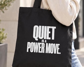 May include: A black tote bag featuring the white text "QUIET IS A POWER MOVE." printed in a bold, sans-serif font on the front. The bag has two black fabric shoulder straps and a rectangular shape, constructed from a solid black textile material.