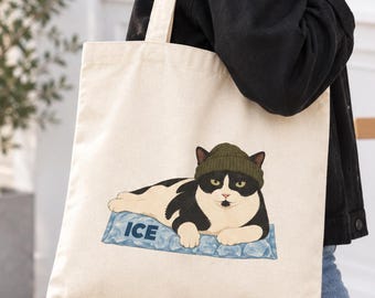 May include: A natural-colored canvas tote bag featuring a graphic illustration of a black and white cat wearing an olive green beanie. The cat is lying down on a rectangular block of blue ice with the word ICE printed in dark blue capital letters.