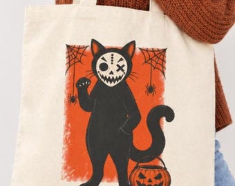 May include: A cream-colored canvas tote bag featuring a graphic illustration of a black cat wearing a white skull mask. The cat stands against an orange rectangular background with spiderwebs and spiders, holding a carved orange jack-o'-lantern bucket.