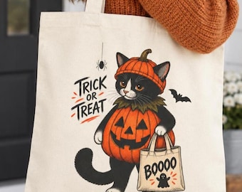 May include: A cream-colored canvas tote bag featuring a black and white cat wearing a pumpkin costume and hat. The cat holds a small bag labeled BOOOO with a ghost icon. The text TRICK OR TREAT appears in black and orange, with a small spider above.