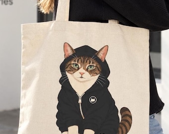May include: A beige canvas tote bag featuring a graphic illustration of a brown and white tabby cat wearing a black zip-up hoodie. The cat has green eyes and a striped tail, and the hoodie includes a small circular logo on the chest area.