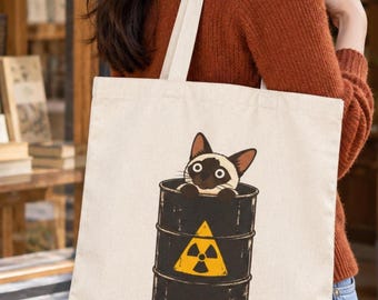 May include: A cream-colored canvas tote bag featuring a graphic illustration of a Siamese cat peeking out from inside a black barrel. The barrel is decorated with a prominent yellow triangular radiation warning symbol on its front surface.