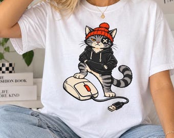 May include: A white t-shirt featuring a graphic of a gray tabby cat wearing a black hoodie and an orange beanie. The cat has an 'X' over one eye and is standing with one paw on a computer mouse with a visible USB cable and connector.