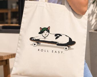 May include: A cream-colored canvas tote bag featuring a graphic illustration of a black and white cat wearing a green baseball cap and a collar, lying on a skateboard. Below the cat, the text reads ROLL EASY. in a simple black font.