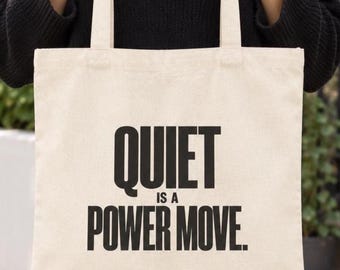 May include: A cream-colored canvas tote bag with two shoulder straps. The front of the bag features bold, black, sans-serif text that reads: QUIET IS A POWER MOVE. The bag is constructed from a woven fabric material and has a rectangular shape.