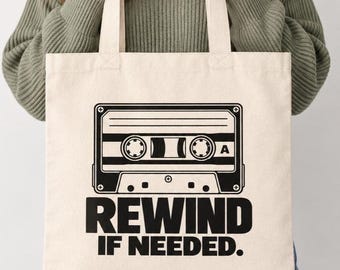 May include: A cream-colored canvas tote bag featuring a black graphic of a vintage cassette tape centered on the front. Below the cassette illustration, the text reads REWIND IF NEEDED in bold, black, capital letters. The bag has two fabric handles.