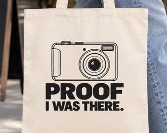 May include: A cream-colored canvas tote bag featuring a black line-art illustration of a digital camera centered above the bold, black, sans-serif text that reads, "PROOF I WAS THERE." The bag has two matching fabric handles and a rectangular shape.