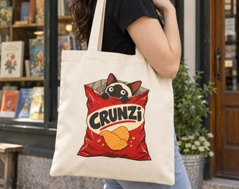 May include: A cream-colored canvas tote bag featuring a graphic print of a Siamese cat peeking out from a red bag of potato chips labeled CRUNZi. The illustration includes two golden-brown ridged potato chips at the bottom of the bag.