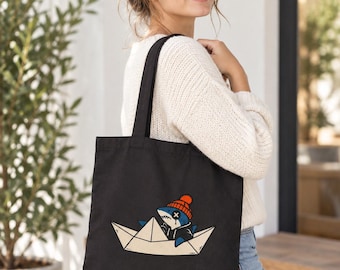 May include: A black tote bag featuring a graphic print of a blue shark wearing an orange beanie and a black hoodie, sitting inside a folded paper boat. The shark has an 'X' over one eye and is depicted in a cartoon style.