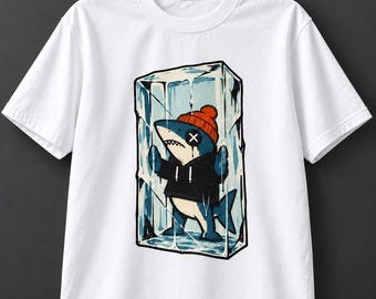 May include: A white short-sleeved t-shirt featuring a graphic print of a cartoon shark wearing an orange beanie and a black hoodie, trapped inside a large block of ice. The shark has an 'X' over one eye and is pressing its fins against the ice.