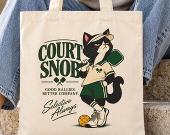 May include: A cream-colored canvas tote bag featuring a graphic of a black and white cat wearing a visor, polo shirt, and shorts. The text reads COURT SNOB, GOOD RALLIES. BETTER COMPANY. Selective Always. The cat holds a paddle near a yellow pickleball.