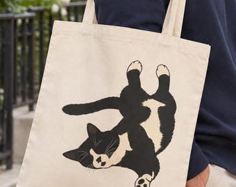 May include: A beige canvas tote bag featuring a graphic illustration of a black and white tuxedo cat lying on its back. The cat is positioned centrally with its paws extended upward and its head turned to the side, showcasing its white chest and face.
