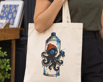 May include: A beige canvas tote bag featuring a graphic print of a black octopus wearing an orange beanie. The octopus is positioned in front of a blue and white spray paint can, holding a smaller blue spray can with one of its tentacles.