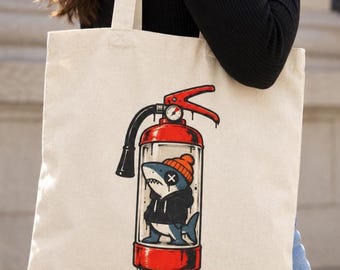 May include: A beige canvas tote bag featuring a graphic print of a red fire extinguisher. Inside the transparent body of the extinguisher, a cartoon shark wearing an orange beanie and a black hoodie is visible, with an X over one eye.
