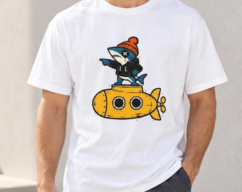 May include: A white short-sleeved t-shirt featuring a graphic print of a cartoon shark wearing an orange beanie and a black hoodie, standing on top of a yellow submarine. The shark is pointing to the left with its right fin.