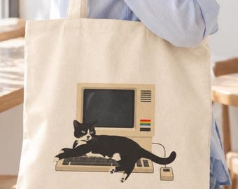 May include: A beige canvas tote bag featuring a graphic illustration of a black and white tuxedo cat lying across a vintage beige computer keyboard and monitor. The computer display is dark, and a small computer mouse is connected by a thin cord.