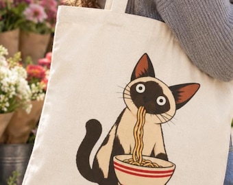 May include: A cream-colored canvas tote bag featuring a graphic illustration of a Siamese cat eating a bowl of noodles. The cat has dark brown ears, face, and tail, and is shown with wide eyes while slurping a long strand of yellow noodles.
