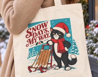 May include: A beige canvas tote bag featuring a graphic of a black and white cat wearing a red Santa hat and scarf, standing next to a wooden sled. The text reads "SNOW DAYS. MY WAY." with a small holly illustration on a blue background.