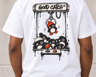 May include: A white t-shirt featuring a graphic on the back of a claw machine game. The illustration shows a claw lifting a small black penguin wearing an orange beanie, with other penguins below. The text GOOD CATCH is printed at the top.