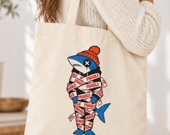 May include: A beige canvas tote bag featuring a graphic of a blue shark wearing an orange knit cap and a black hoodie. The shark is wrapped in white packing tape printed with the red text FRAGILE. The shark has an X over one eye.