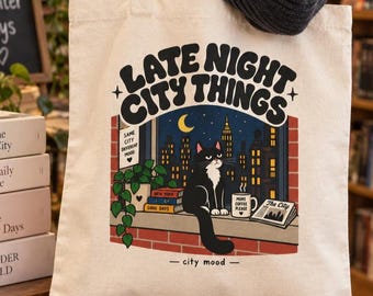 May include: A beige canvas tote bag featuring a graphic illustration of a black and white cat sitting on a windowsill at night. The text reads LATE NIGHT CITY THINGS, SAME CITY DIFFERENT MOOD, NEW YORK, GOOD DAYS, MORE COFFEE PLEASE, and The City.