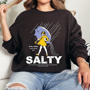 May include: A dark brown sweatshirt featuring a graphic of a person under a blue umbrella, with the word "SALTY" below. The graphic includes the text "All the flavors and you choose to be".