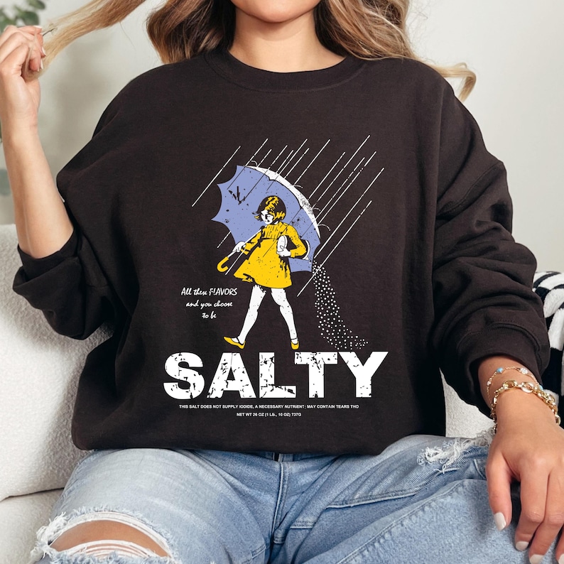 May include: A dark brown sweatshirt featuring a graphic of a person under a blue umbrella, with the word "SALTY" below. The graphic includes the text "All the flavors and you choose to be".