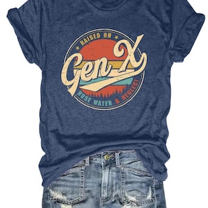 Puede incluir: Camiseta azul marino con un dise&ntilde;o retro que dice "Raised on Gen X, Hose Water & Neglect" en un dise&ntilde;o circular con acentos naranja, amarillo y turquesa. La camiseta se combina con pantalones cortos de mezclilla.