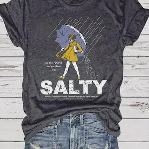 May include: Dark gray t-shirt with a graphic of a person under an umbrella, with the word "SALTY" in large white letters. The person is wearing a yellow coat and white pants. The text "All these flavors and you choose to be" is also on the shirt.