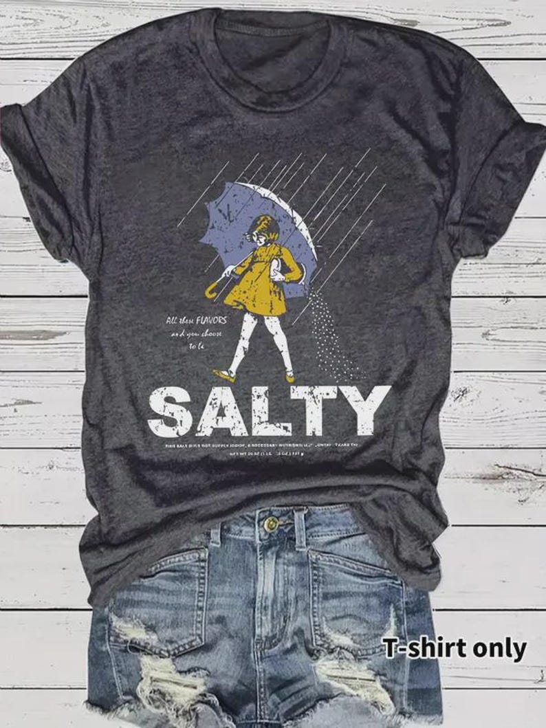 May include: Dark gray t-shirt with a graphic of a person under an umbrella, with the word "SALTY" in large white letters. The person is wearing a yellow coat and white pants. The text "All these flavors and you choose to be" is also on the shirt.