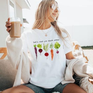 May include: White t-shirt with the text "Let's root for each other" and a watercolor illustration of various vegetables. The vegetables include a purple carrot, a red beet, a green leafy vegetable, an orange carrot, and a red beet.