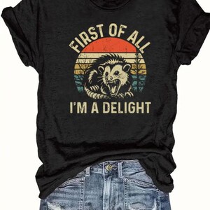 May include: Black t-shirt featuring a graphic of an opossum inside a circular design with a retro sunset. The text reads "FIRST OF ALL I'M A DELIGHT". The shirt is paired with denim shorts.