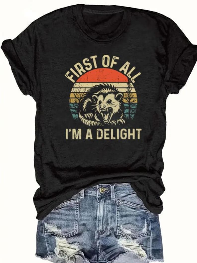 May include: Black t-shirt featuring a graphic of an opossum inside a circular design with a retro sunset. The text reads "FIRST OF ALL I'M A DELIGHT". The shirt is paired with denim shorts.
