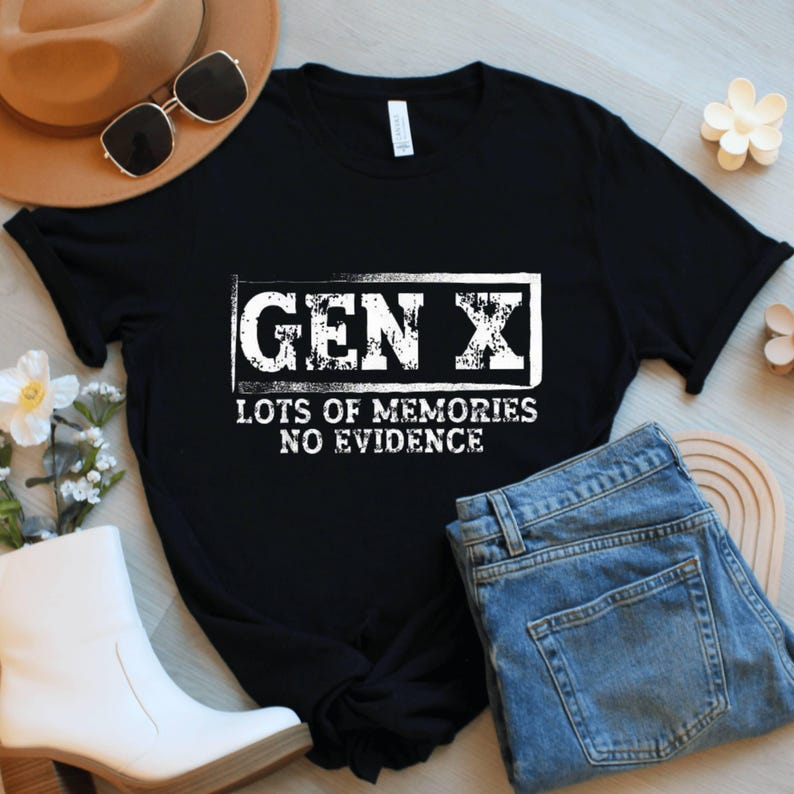 May include: Black t-shirt with the text "GEN X LOTS OF MEMORIES NO EVIDENCE" in a white, distressed font. The shirt is displayed with blue jeans, a brown hat, sunglasses, and white boots.