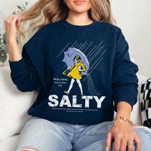 May include: Navy blue sweatshirt with a graphic of a girl in a yellow dress holding a purple umbrella in the rain. The word "SALTY" is printed in large white letters below the illustration. The text "All then flavors and you choose to be" is above the illustration.